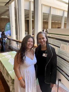 Researchers Samia Dutra and Frankie Hale stand in front of a poster at the state capitol. Samia is wearing a white dress, and Frankie is on her right, wearing black.