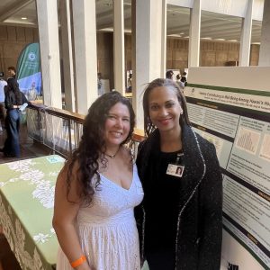 Researchers Samia Dutra and Frankie Hale stand in front of a poster at the state capitol. Samia is wearing a white dress, and Frankie is on her right, wearing black.